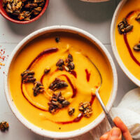 Holding a spoon in a bowl of carrot ginger soup topped with sriracha and spicy seeds