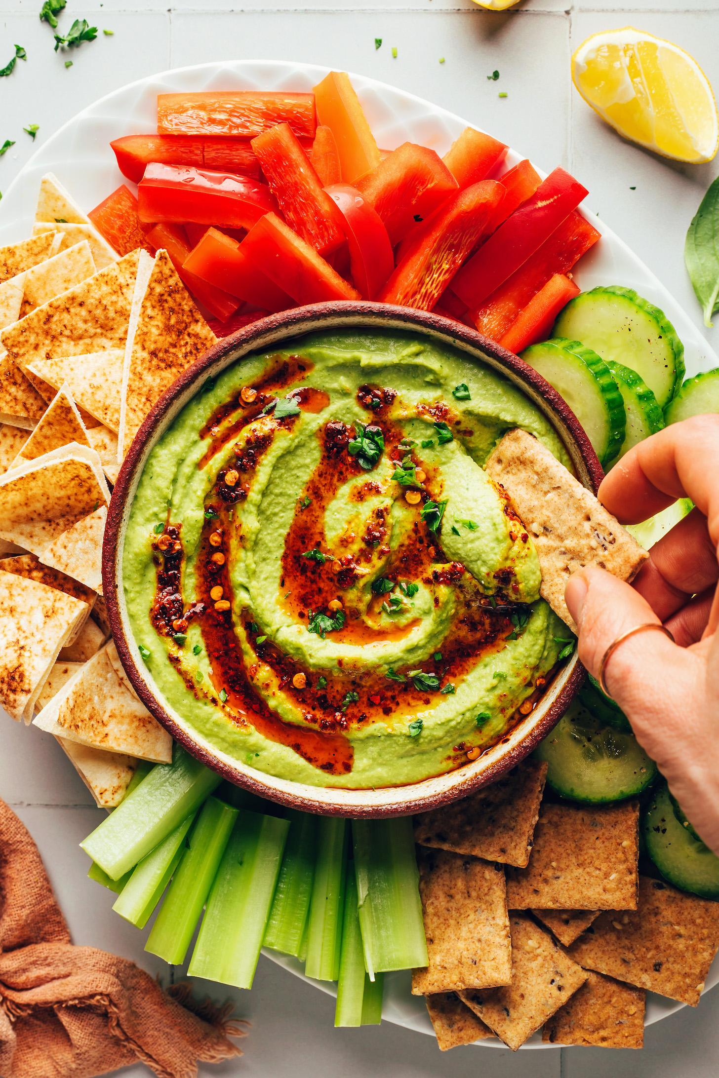 Dipping a cracker into a bowl of green goddess hummus topped with chili oil and parsley