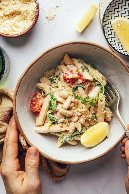 Hands holding a bowl of lemon garlic pasta with white beans and arugula