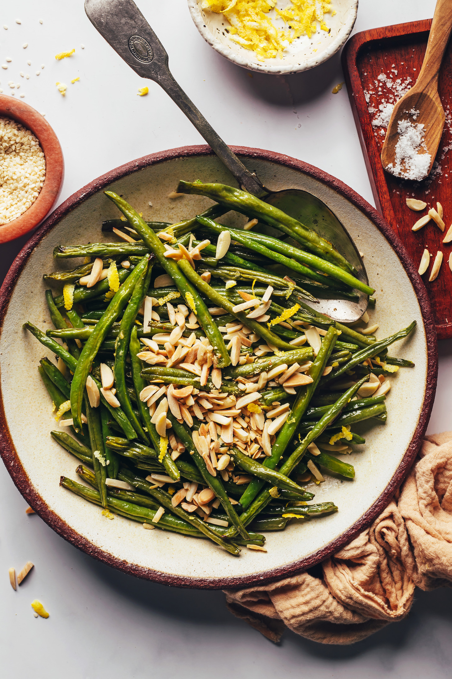 Lemon zest, sesame seeds, salt, and slivered almonds next to a bowl of roasted green beans topped with lemon zest and almonds