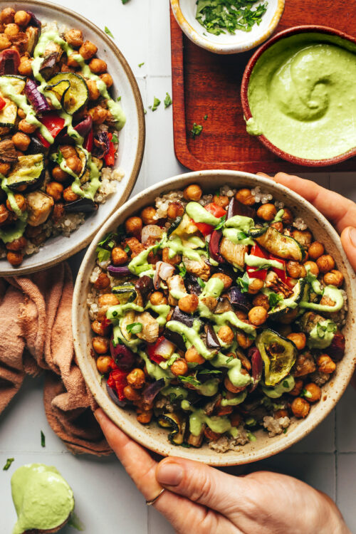 Hands holding the sides of a bowl of roasted chickpeas, quinoa, and veggies topped with green tahini sauce