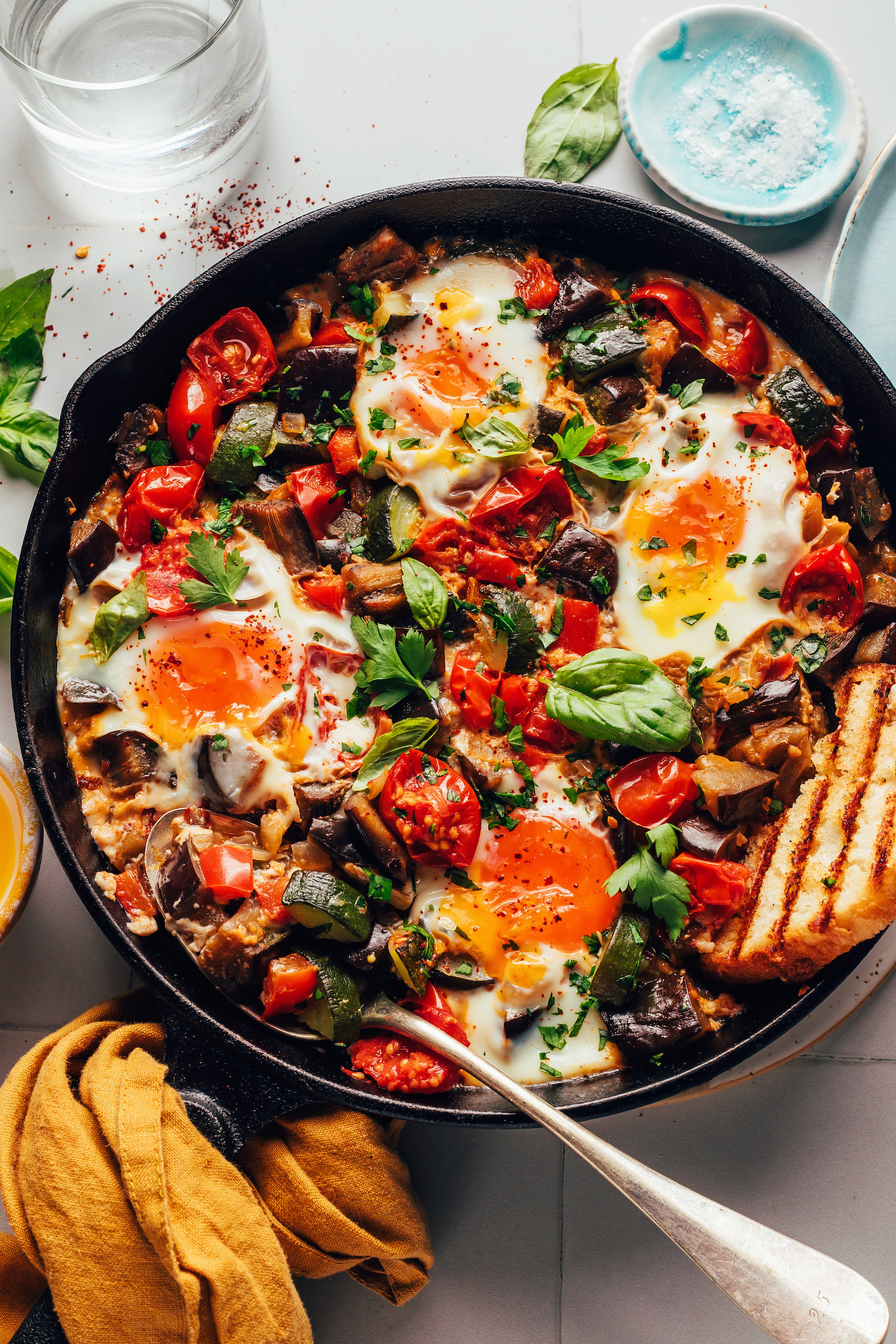 Spoon and slice of toasted bread resting in a pan of skillet ratatouille and eggs