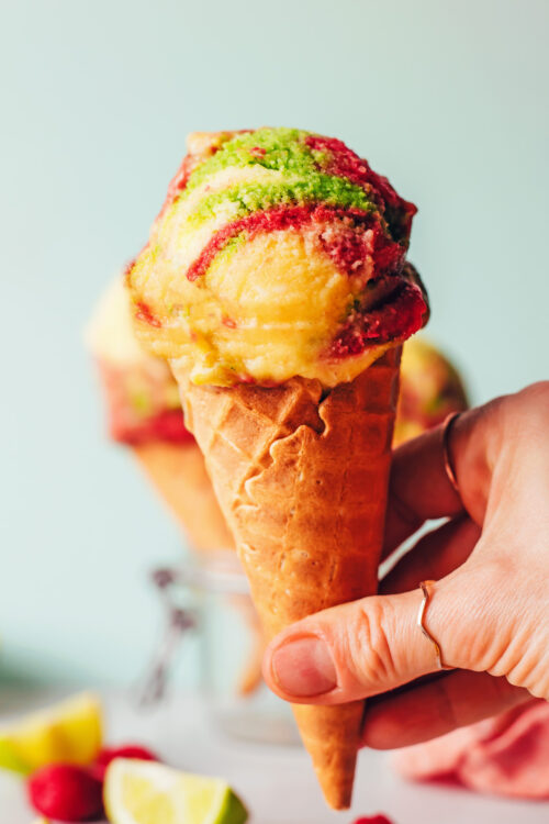 Hand holding a cone with a scoop of vegan rainbow sherbet ice cream on top