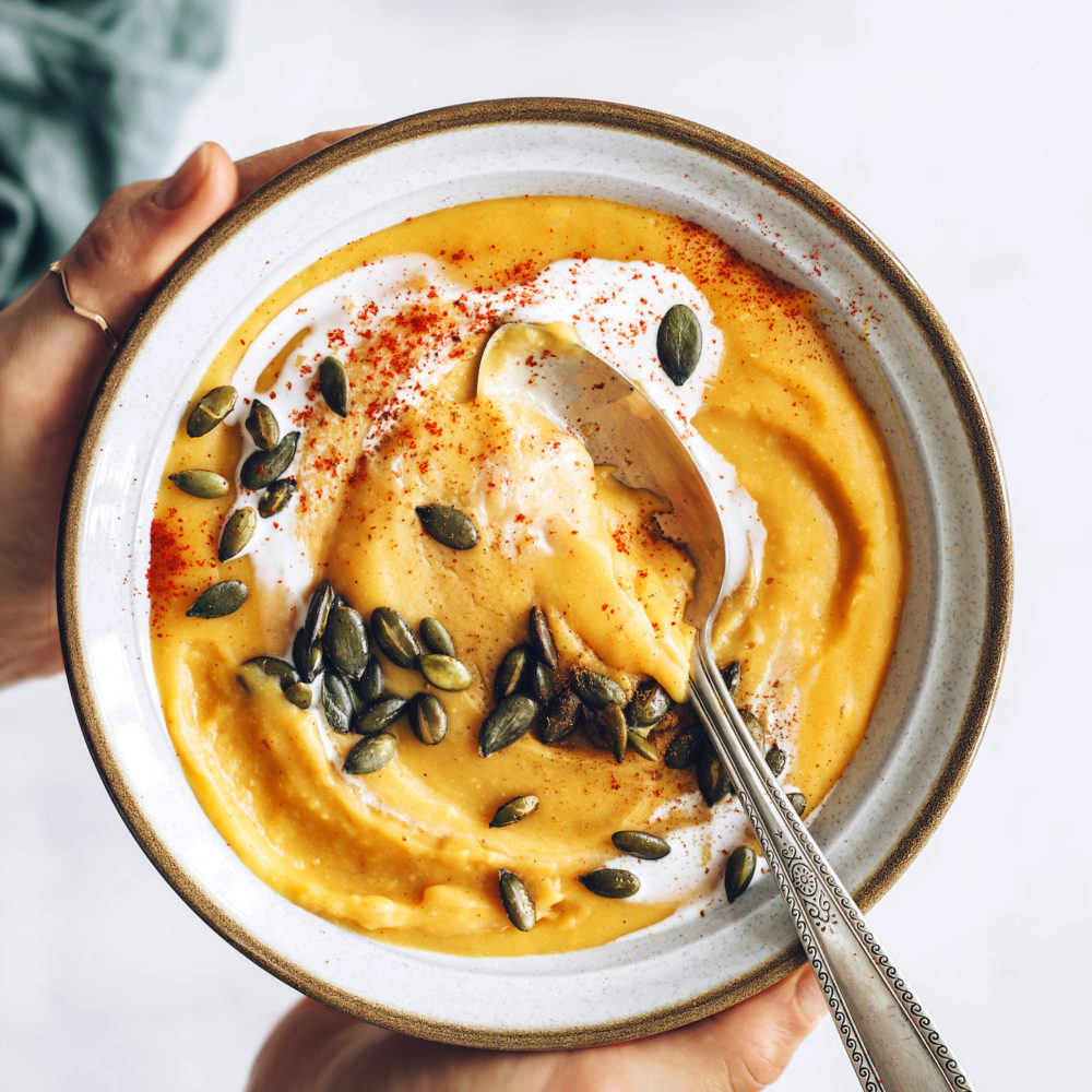 Holding a bowl of creamy sweet potato lentil soup topped with coconut milk and pepitas